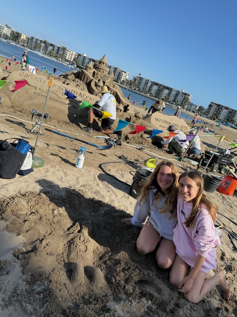 two girls sitting on sand next to a sandcastle