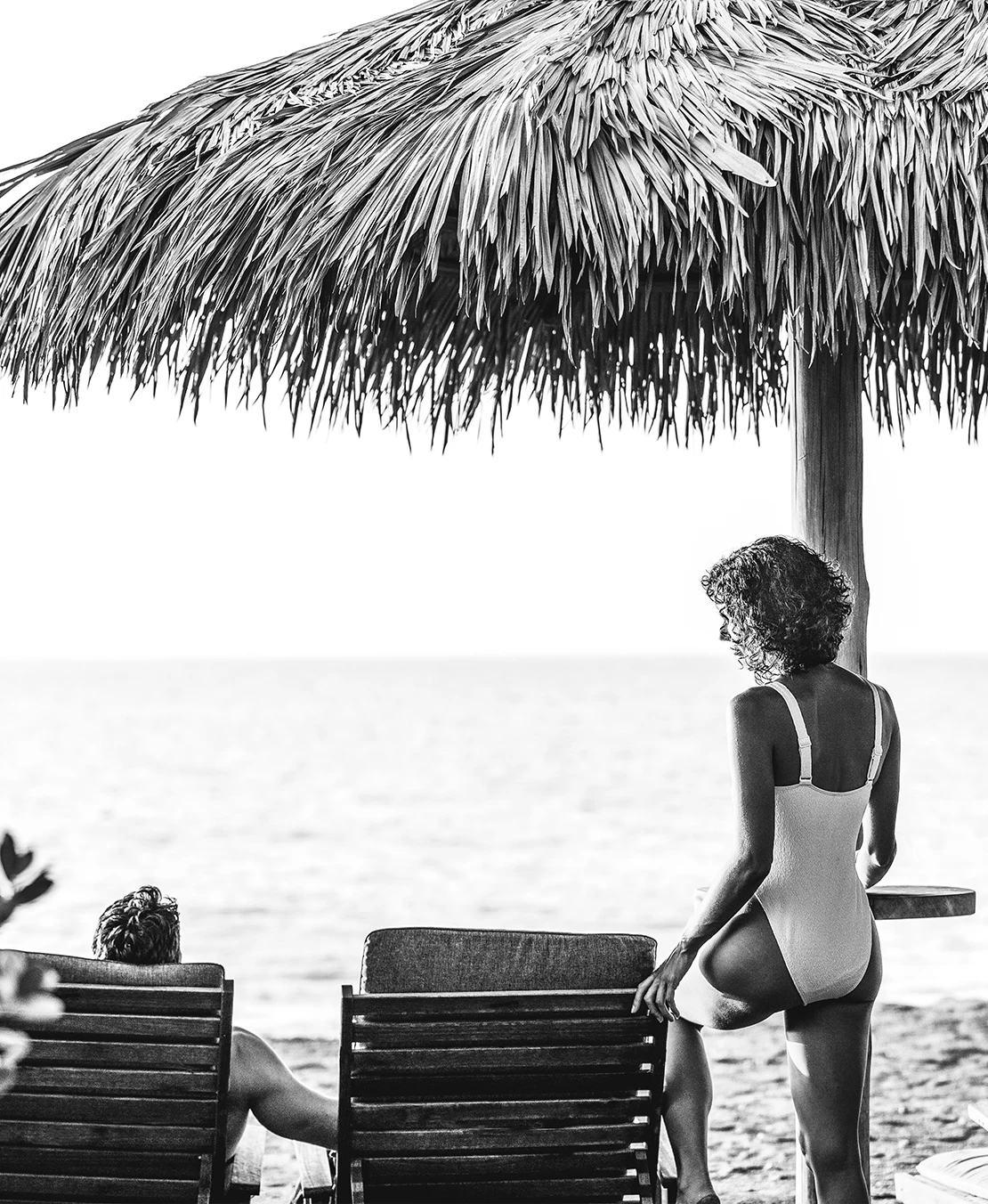 a woman sitting on a chair under a palm tree umbrella on a beach
