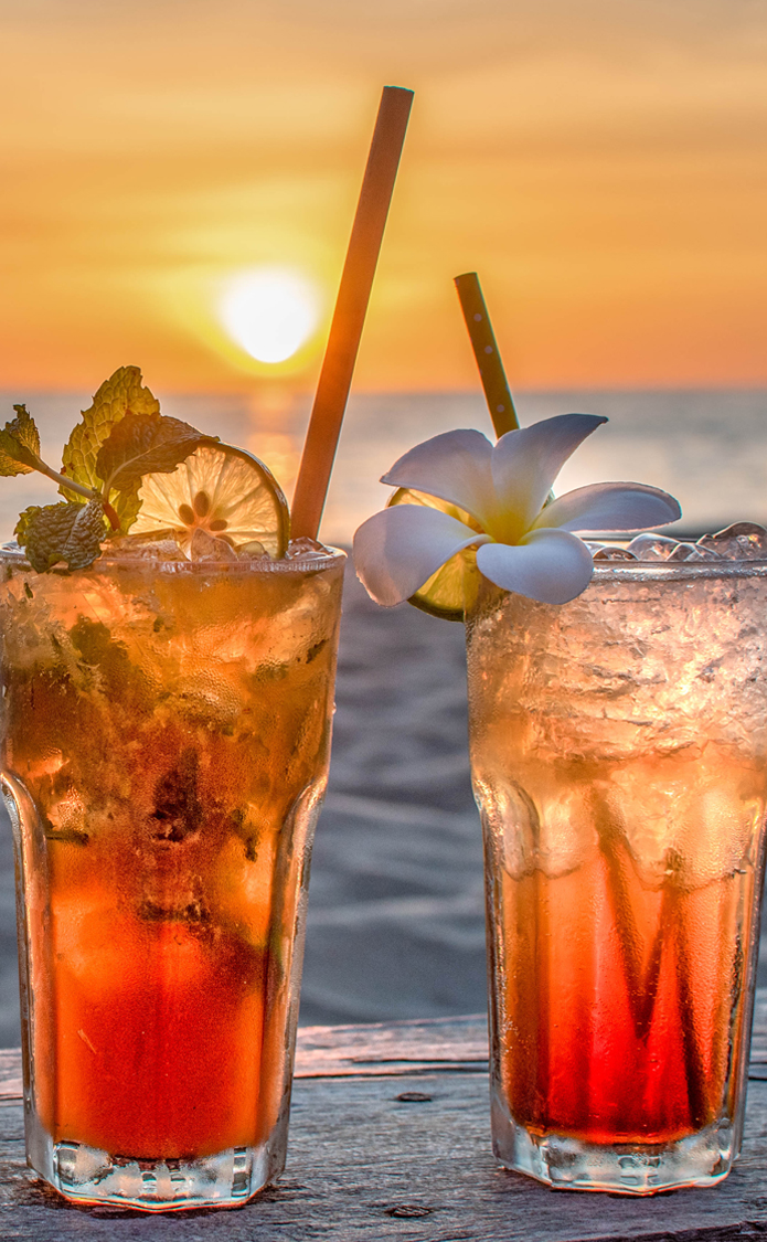 two glasses with a red drink and flower decals and a straw with the ocean and sunset in the background