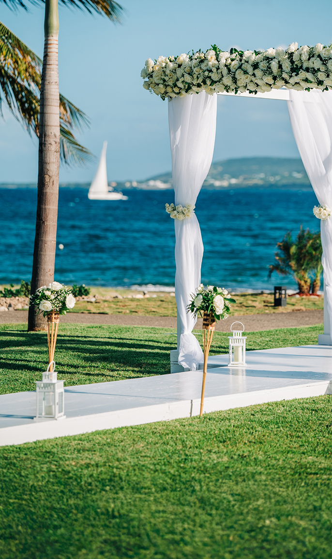 a white wedding arch with white flowers and candles on a lawn with a sailboat in the background