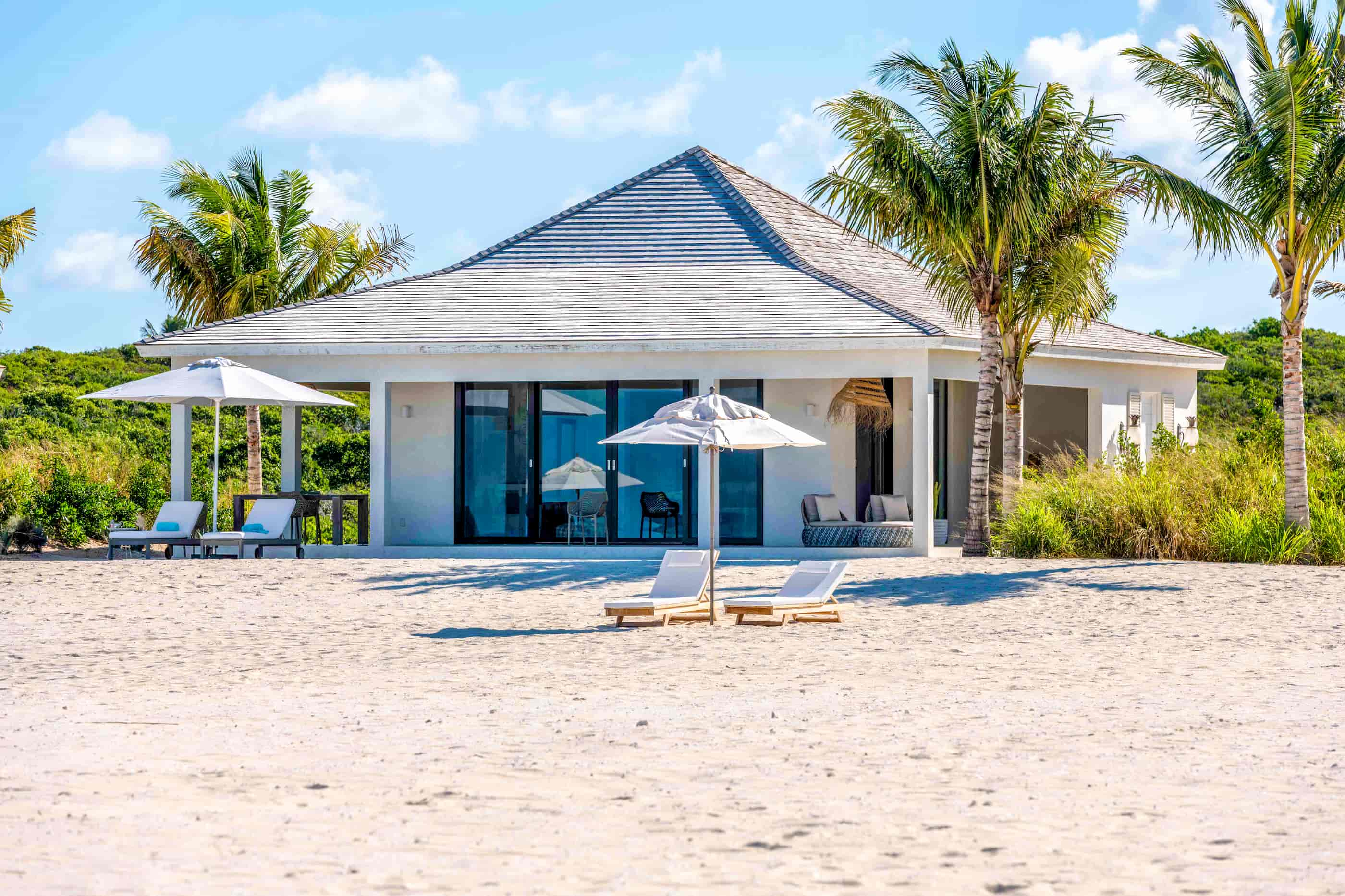 a white house with lounge chairs and umbrellas on a sandy beach
