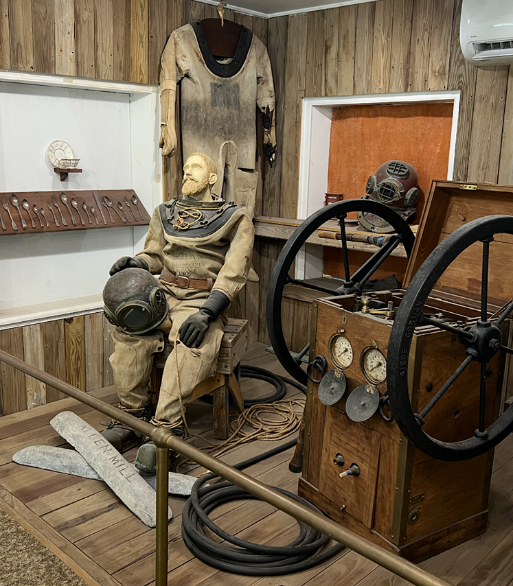 a man in a suit sitting in a chair with old diving helmet and equipment